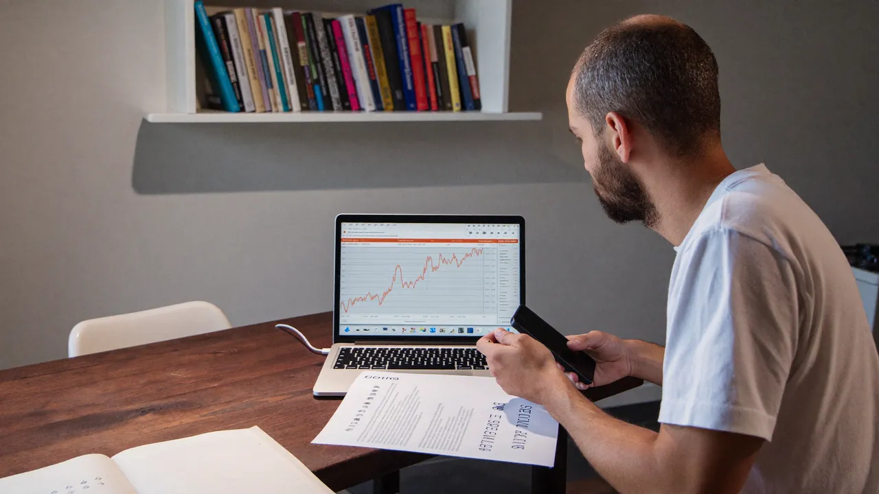 A man at a kitchen table looks at a chart on his laptop, a sheet of figures beside it.