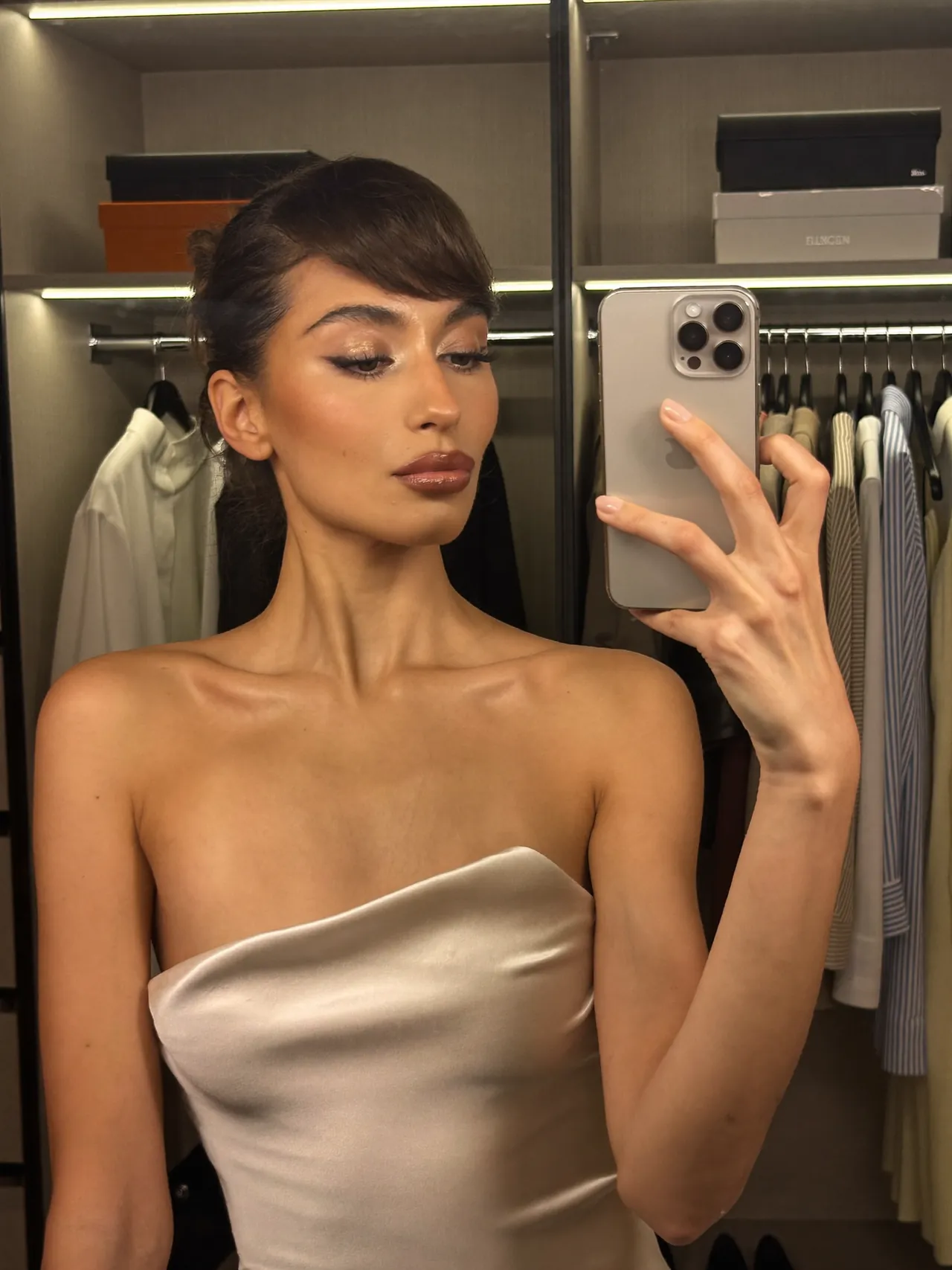 A young woman films herself in her bedroom mirror with her phone, daylight behind her.